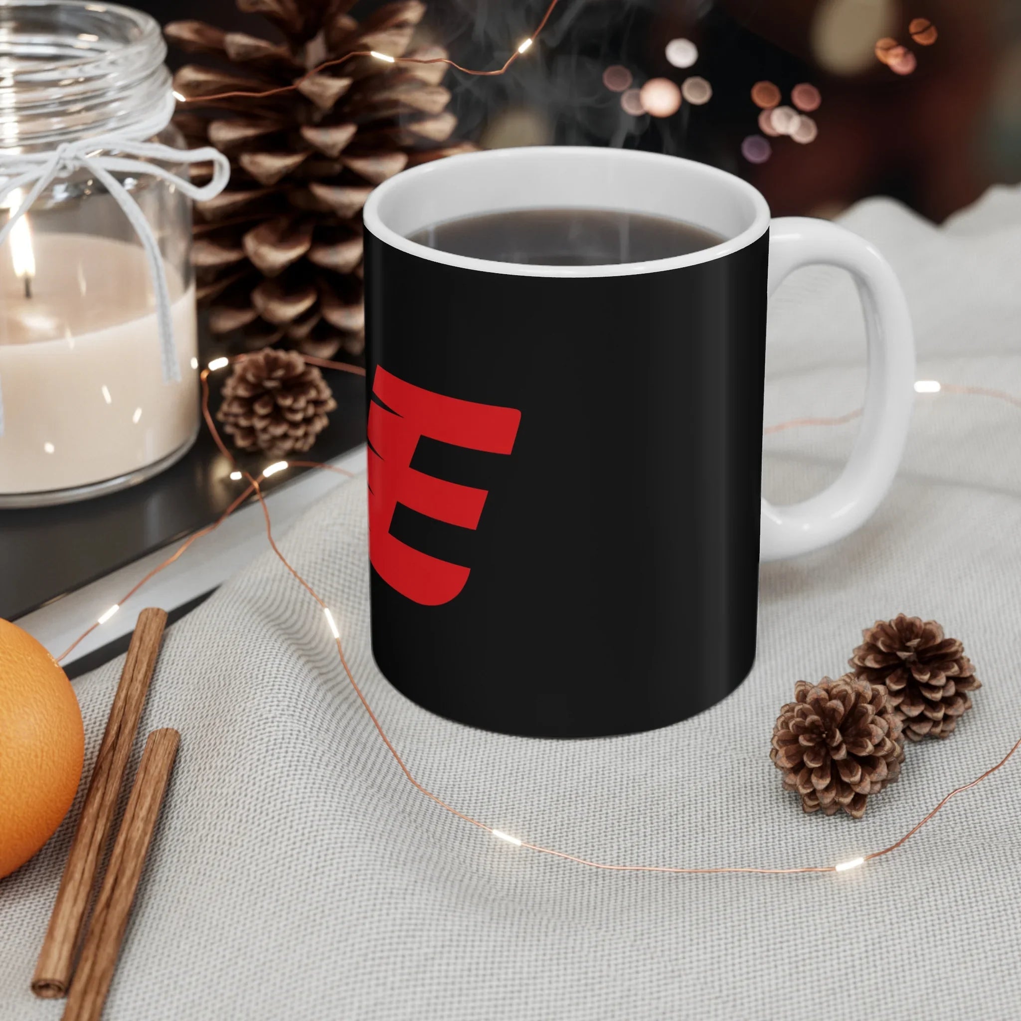 Black mug with red graphic letter E, surrounded by cozy winter decor, pinecones, candle, and coffee steam